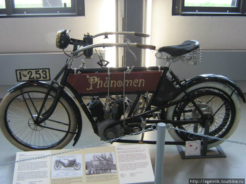 Phänomen red 1905
Велосипедная фабрика «Phanomen», основанная еще в 1888 году, свой первый мотоцикл изготовила в начале XX века. Типичные для своего времени мотоциклы оснащались одно- и двухцилиндровыми V-образными двигателями «Фафнир» с автоматическим впускным клапаном. Привод заднего колеса был ременным. С1907 года в серию пошли трехколесные мотоциклы в пассажирском и грузовом вариантах, а к 1910 году автомобили полностью вытеснили двухколесную технику из производственной программы.

Чистокровный «немец» — мотоцикл Phanomen 1905 года. 500-кубовая «двойка» Fafnir мощностью около 2 л.с. разгоняла машину до скорости 75 км/ч. Для освещения служил ацетиленовый фонарь, газ для него вырабатывался в «генераторе» позади фары. Обратите внимание на обилие рычажков на бензобаке — на них выведено управление и карбюратором, и зажиганием. Мюнхен, Германия