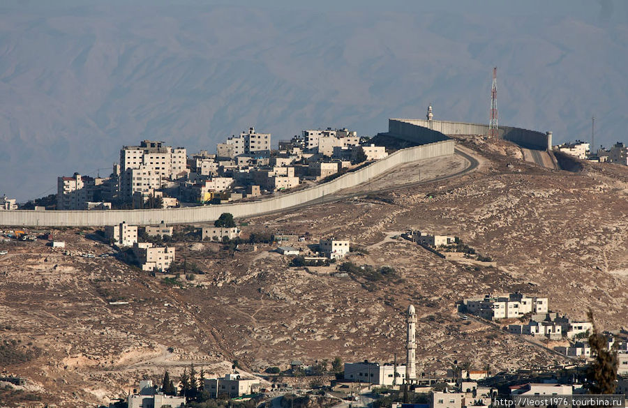 Та самая заградительная стена, которую Израиль начал строить для отгорождения Израиля от Западного берега реки Иордан в 2003 году и строит до сих пор, чтобы защититься от нападений палестинцев и которая вызывала много споров, в частности из-за того что Израиль отошел от т.н. зеленой линии, по которой проходит граница.
Общая длинна более 700км, 10% представляет собой бетонную стену как на снимке, остальное забор с колючкой с 60 метровой зоной отчуждения — на Берлинскую стену смахивает.
По статистике после строительства количество терактов и нападений снизилось. Иерусалим, Израиль