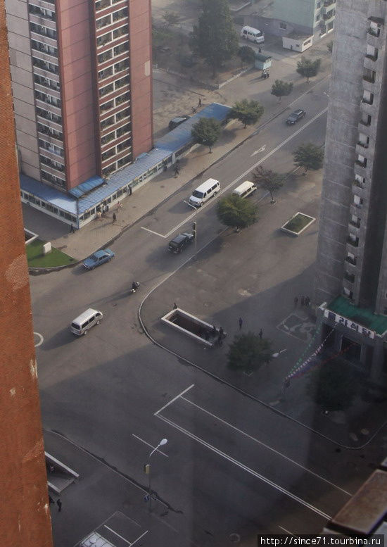 3. Самое потрясающее зрелище в Пхеньяне в семь утра, тем более после Пекина, это пустые улицы и строгие соблюдения правил движения. При всём неналичии другого транспорта, машины останавливаются  на красный свет светофора, мигают нужными поворотниками, скорость соблюдают наистрожайше. Хотя даже не это потрясает, а то как тщательно водители  стараются остановиться в рамках расчерченных белых линий, как аккуратно двигают с места и поворачивают.
Ещё больше чем поведение водителей, потрясает поведение пешехрдов. На этом  перекрёстке нет наземного перехода, есть подземный. Быстренько через улицу эдесь никто не перебегает, даже если что-то надо прямо напротив, а до перехода далеко. Здесь все дисциплинированно доходят до перехода, переходят на другую сторону и идут в продолжают путь в нужном направлении, даже если надо возвращаться обратно.
Низкая  пристройка под крышей — магазин, где время от времени наблюдались очереди. КНДР